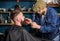 Barber busy with grooming mustache of hipster client, beauty supplies on background. Hipster with beard and mustache