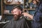 Barber with clipper trimming hair on nape of client. Barber with hair clipper works on haircut of bearded guy barbershop