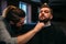 Barber combing beard of client man in salon cape