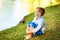 Barefoot 5 years old boy sit on lake shore and look away. Child in white shirt and blue jeans without shoes near water close to