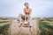 Barefoot brunette in linen pants and bare shoulders sitting on a hay bales in warm autumn day. Woman looking at camera. Behind her