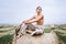 Barefoot brunette in linen pants and bare shoulders sitting on a hay bales in warm autumn day. Woman looking at camera. Behind her