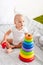 Barefoot child in white clothes playing with toy pyramid on bed