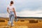 Barefoot girl with sneakers in hand stands on a haystack on a bale in the agricultural field after harvesting