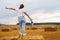 Barefoot girl with sneakers in hand stands on a haystack on a bale in the agricultural field after harvesting