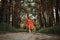 Barefoot happy young woman in red dress with hand raised dancing in pine forest at summer day.