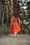 Barefoot happy young woman in red dress with hand raised dancing in pine forest at summer day.