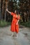 Barefoot happy young woman in red dress with hand raised dancing in pine forest at summer day.