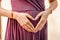 Barely showing young expecting mother wearing a pleated purple dress while forming a heart with her hands over the belly