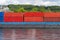 A barge carrying many containers on the Rhine in western Germany, trees and buildings in the background.