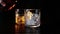Barman pouring whiskey in the two glasses with ice cubes on wood table and black dark background, focus on ice cubes