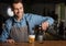 Barman pours beer from bottle in glass, standing behind bar counter in interior