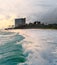 A barreling wave crashes and creates a leading line into beautiful scenic sunset and tall buildings on the coast. Silhouetted palm