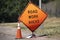 Battered Road Work Ahead sign and traffic cone sitting by road with blurred background