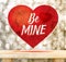 Be mine word in red heart floating above wood table at gold blur