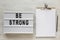 `Be strong` words on a lightbox, noticepad with blank sheet of paper on a white wooden background, top view. Overhead, from abov