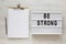 `Be strong` words on a lightbox, noticepad with blank sheet of paper on a white wooden surface, top view. Overhead, from above,