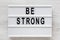 `Be strong` words on a lightbox on a white wooden surface, top view. Overhead, from above, flat lay