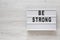 `Be strong` words on a lightbox on a white wooden surface, top view. Overhead, from above, flat lay. Copy space