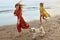 Beach. Girls With Dog Walking Barefoot On Sandy Coast. Fashion Women In Bohemian Clothing With Pet