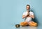 Bearded aged man in dhoti clothes. Praying, sitting on floor in lotus pose on blue background. Singing bowl, rosary nearby