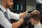 Bearded barber smiling and trimming haircut of his male client.