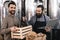 Bearded brewer in apron with laptop takes out bottle from box, which is held by cheerful man.