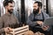 Bearded brewer in apron looks at merry man holding box of empty bottles.