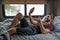 A bearded brown haired hypster man wearing a red t-shirt lays down laughing relaxing on his bed in his camper talking to someone