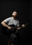 Bearded brutal guitarist plays an acoustic guitar in a black room and looking in the camera. The concept of music