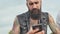 Bearded and brutal man with tattoos looks into the phone on the street.