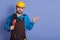 Bearded Caucasian builder with grinder in hands posing isolated over blue background and pointing index finger aside at copy space