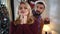 Bearded Caucasian man in Christmas hat coming to pretty blond woman from background, closing her eyes and giving gift