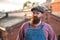 Bearded craftsman wearing blue overalls, checked shirt and cap in vintage style, looking at camera, outdoors. Family workshop, old