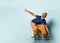 Bearded elderly man in t-shirt, sunglasses, orange pants, hat, gumshoes. Riding black skateboard, posing on blue background