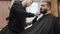 A bearded hipster Barber dries and styles a client's beard in a barbershop with a hair dryer and comb. In the