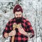 Bearded hipster checks ax blade in winter forest. Lumberjack works in snowy day. Bearded guy wears checkered shirt and knitted hat