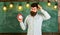 Bearded hipster holds clock, chalkboard on background, copy space. Man with beard and mustache on forgetful face itching