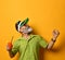 Bearded male in green sun visor, shirt and sunglasses. Holding glass of squeezed juice, clenched fist, posing on orange background