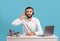 Bearded male helpline operator with headphones and laptop answering customer's call at desk over blue studio background