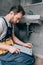 Bearded male plumber with toolbelt writing in clipboard near broken sink