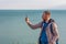 Bearded man in black raincoat on a blue sea landscape background with smartphone in hands takes seascape photography