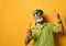 Bearded man in green sun visor, shirt and sunglasses. Holding glass of squeezed juice, clenched fist, posing on orange background