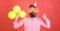 Bearded man showing thumb up gesture. Man with trendy beard blowing party whistle. Hipster with colorful balloons on red