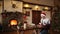 Bearded man sitting and tying bow on a gifts near fireplace, wearing Christmas hat wrapping boxes in paper, decorating