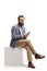 Bearded man sitting on a white cube and holding a smartphone