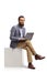 Bearded man sitting on a white cube and using a laptop computer