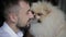 Bearded man in white shirt holds his white fluffy spitz in hands and tries to rub his nose with him, close view. Cute