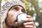 Bearded man in wool knitted hat intently drinks hot tea or coffee from mug, side view on face, background of forest