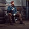 A bearded mechanic dressed in a uniform, sits on old car tires in the garage.